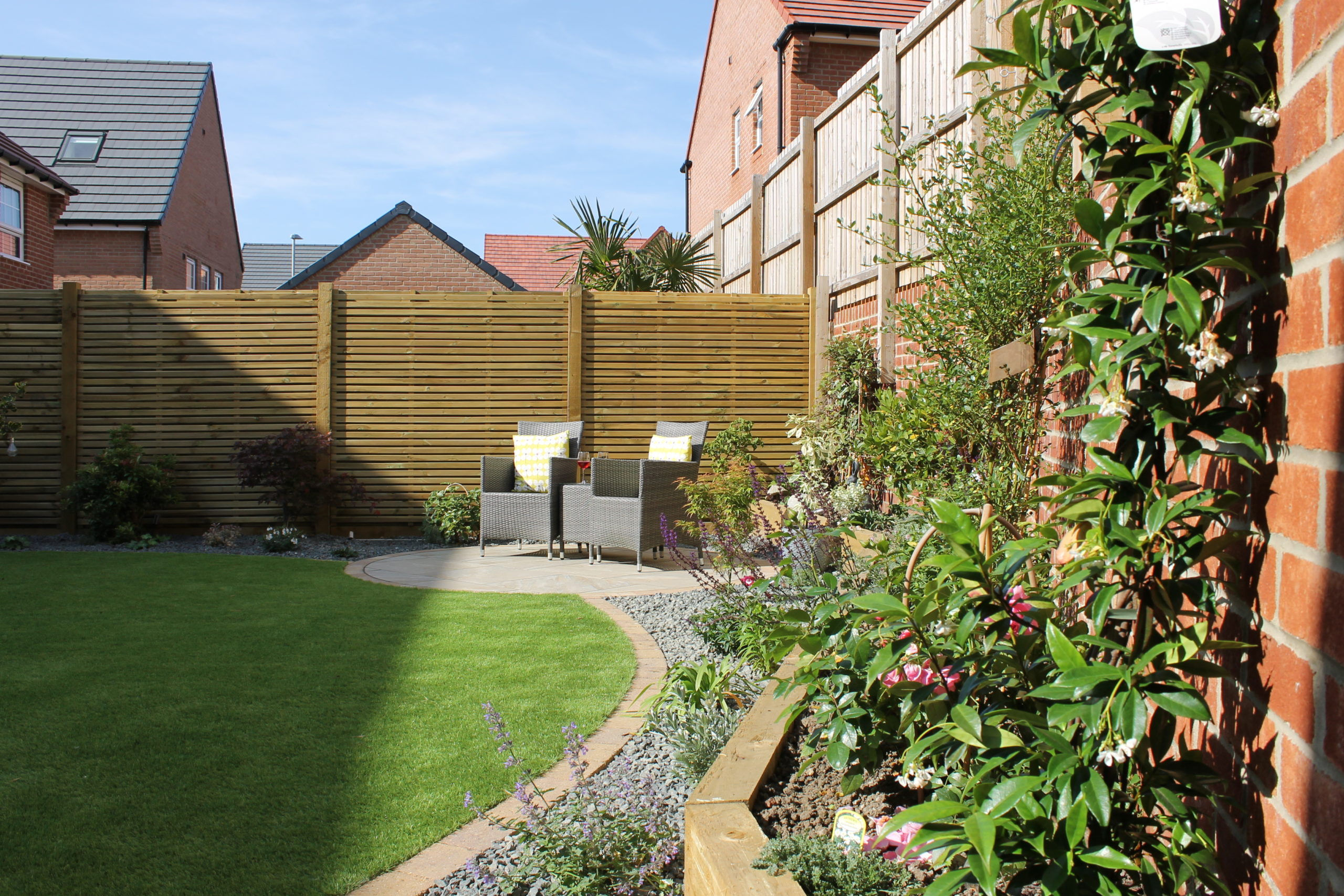 Corner of a garden with a brown fence, plants and grey chairs